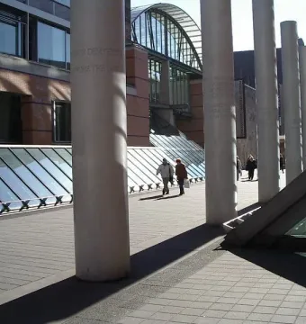 Way of Human Rights pillars lining a paved walkway beside modern buildings, with people strolling in sunlight.