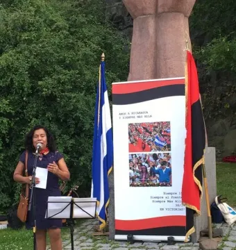 A woman in a blue dress speaks into a microphone near La Mano, with flags and a banner behind her.