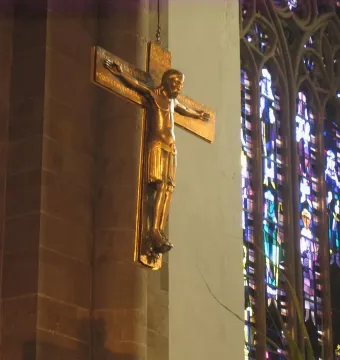 Mindener Cross hanging on a wall beside a stained-glass window in a dimly lit interior.