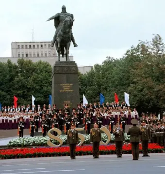 Monument to Yuri Dolgorukiy on a dark pedestal, with a crowd, choir, and band gathered in front of it.