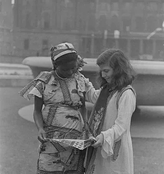 Two women examine a patterned piece of fabric or paper together, one wearing a printed dress and headscarf and the other a light outfit with a decorative vest.
