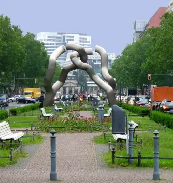 The Berlin sculpture of segmented grey tubes stands in a tree-lined street with cars, benches, and urban buildings nearby.