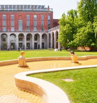 The Mystery Baths fountain with sculpted figures and a colorful swan in front of a red brick building and trees.