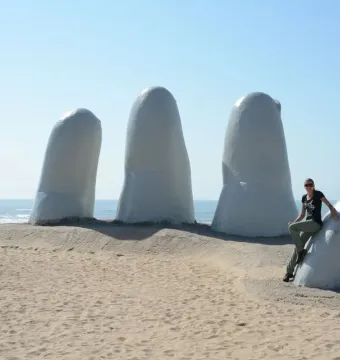 The Hand sculpture rising from a sandy beach with the ocean and blue sky behind it, with a woman seated on one of the fingers.