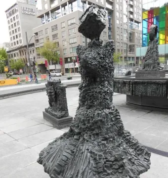 Dark bronze sculptures from La Joute in an urban plaza, with a large abstract figure in the foreground and a fountain base behind it.