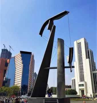 Puerta 1808, a tall angular metallic sculpture, stands in a busy urban plaza with pedestrians, high-rise buildings, and cranes in the background.