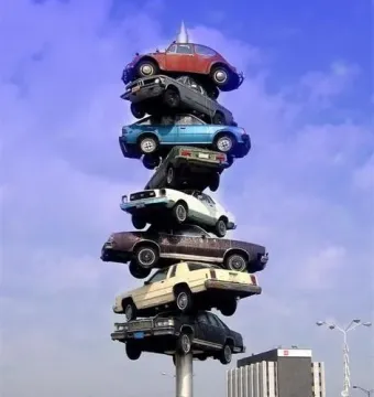 Spindle sculpture with cars stacked vertically on a central pole above a concrete base.