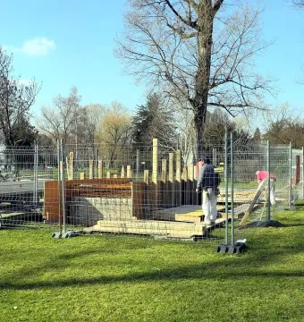 An outdoor construction site in a grassy park, fenced off with temporary metal barriers, with a partly built wooden and rebar structure and a few people nearby.