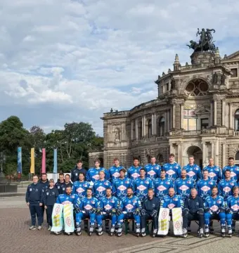 King Johann Memorial standing on a high stone pedestal in a public square, with ice hockey players posing near the opera house in the background.