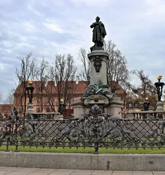 Adam Mickiewicz Monument showing a bronze cloaked figure on an ornate stone pedestal, with bare trees and wrought-iron fencing nearby.