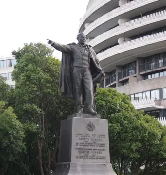 Bronze statue of Benito Juarez on a dark pedestal, pointing forward with a modern building and trees behind it.