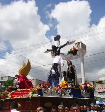 A religious procession carries an ornate float with the San Juan Patrón sculpture and a crucifix, surrounded by flowers and costumed participants.