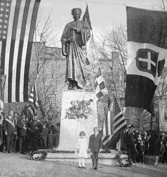 Dante Alighieri statue on a granite pedestal, surrounded by flags and a crowd at a dedication ceremony.