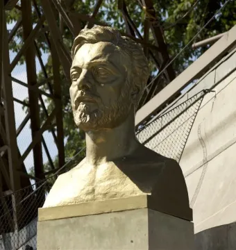 Gustave Eiffel bust on a pedestal with the Eiffel Tower structure in the background.