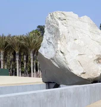 Levitated Mass sculpture with a large boulder appearing to hover above a concrete trough, with palm trees and blue sky behind it.