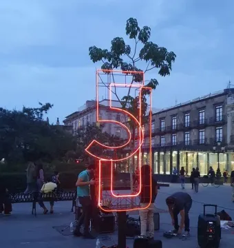 Antimonumento 5J glowing orange in a public plaza at twilight, with people and audio equipment nearby.