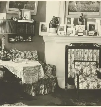 A small dark crouching Venus statuette sits on a crowded mantelpiece beside a clock and framed photographs.