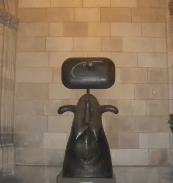 Femme, a dark abstract sculpture with a rounded rectangular head, tapered body, curved arm-like forms, and a teardrop-shaped indentation, stands on a base against a stone wall.