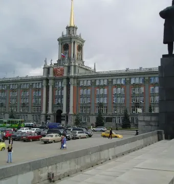 Lenin Monument seen from behind with one arm raised, in front of a large historic building and parked cars.