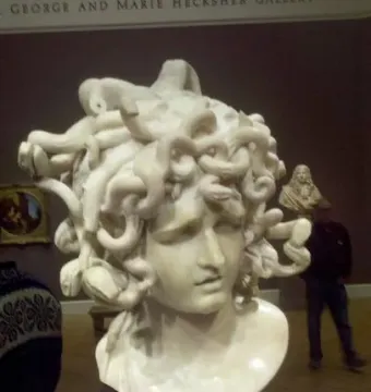 Marble bust of Medusa with snakes carved as hair, displayed on a pedestal in a gallery.