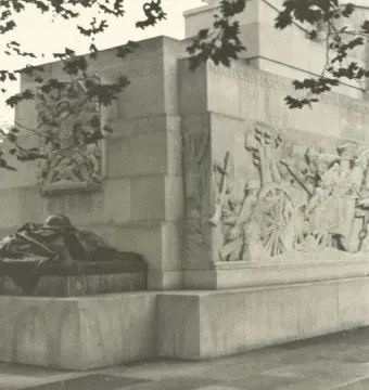 Royal Artillery Memorial with stone relief carvings and a helmet sculpture on a lower platform, partly framed by tree branches.