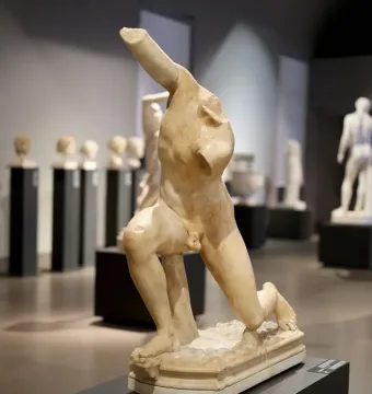 Damaged marble sculpture of the Subiaco Ephebe with missing head, arms, and lower legs, displayed on a pedestal in a museum hall.