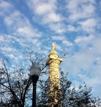 The Washington Monument stone column rises above trees and a streetlamp against a bright blue sky with scattered clouds.