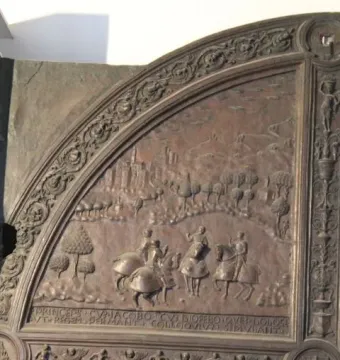 Ornate bronze gate panel with relief scenes of horsemen, figures in a landscape, a Latin inscription, and a crowded battle below.