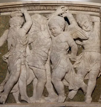 Marble relief from the Pulpit of Prato Cathedral showing dancing figures and winged putti against a dark mosaic background.
