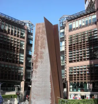 Fulcrum, a tall rusty steel sculpture, stands in an urban plaza beside modern office buildings and hedges.