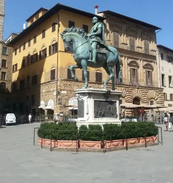 Cosimo I equestrian monument on a tall stone pedestal in a city square, with historic buildings and a few people nearby.