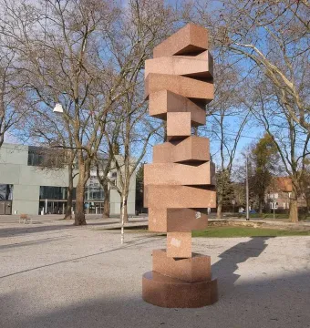 Endlose Treppe by Max Bill, a tall reddish-brown stone sculpture on a paved plaza with trees and a modern building behind it.