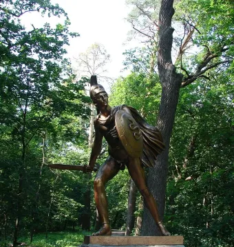 Greek Warrior Monument: a bronze armored warrior with a crested helmet, shield, and blade among tall trees.