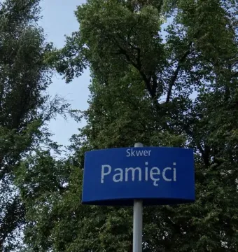 Blue Skwer Pamięci street sign mounted on a light-colored pole with trees and a building in the background.