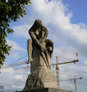 Tauzieher stone sculpture of a crouching figure fastening a thick rope to a post, with cranes and a blue sky behind it.