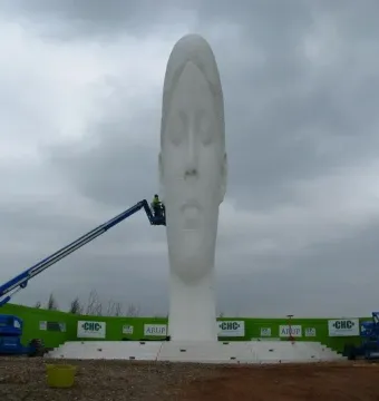 The Dream sculpture stands tall with a serene face under a cloudy sky, while a worker in a blue hydraulic lift works near the base.