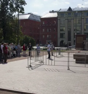The Solovetsky Stone stands on a dark base in a paved square, with people gathered nearby behind metal barriers.