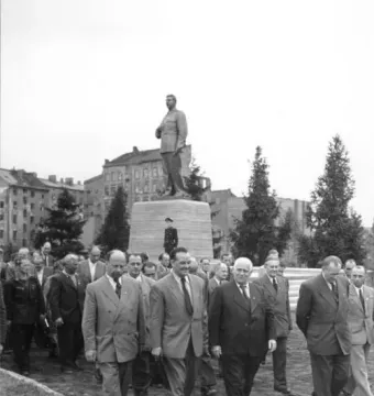 A tall statue of Joseph Stalin stands on a multi-tiered pedestal in a public square, with several men walking in the foreground.