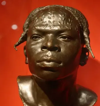 Dark bronze bust of an Aruwimi native with detailed hair or head markings against a red background.