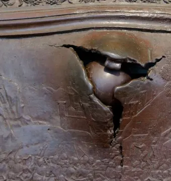 Close-up of the Bronze Gate in Naples, showing a jagged rupture with a rounded object lodged in the bronze panel.