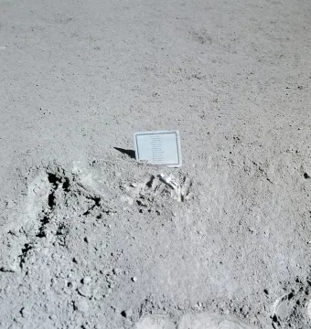 A small white fallen astronaut figure lies beside a plaque on a gray lunar surface.