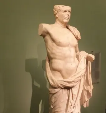 The General of Tivoli, a damaged white marble statue of a male figure with a broken head and missing upper arms, draped with a cloak against a green wall.