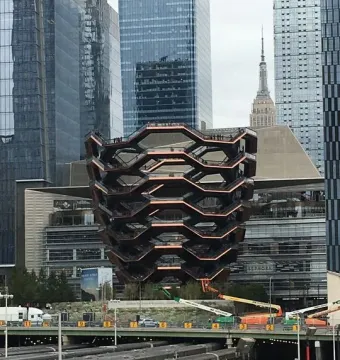 The Vessel sculpture in Manhattan, with honeycomb-like stairways beside glass skyscrapers and train tracks in the foreground.