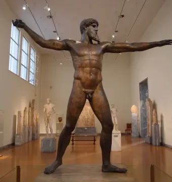 The Artemision Bronze stands on display in a museum gallery, showing a bearded nude figure with both arms outstretched.