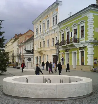 The Capitoline Wolf statue on a tall stone plinth in a European town square, with pedestrians, a circular fountain, and historic buildings nearby.