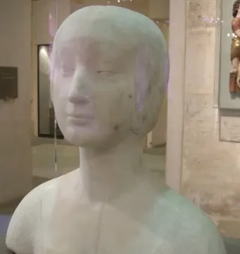 White marble bust of a princess with a serene expression and close-fitting cap on a dark base in a museum display.