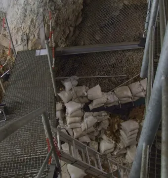 Archaeological excavation site inside the Hohle Fels cave with metal walkways, stairs, and sandbags among rocky walls.