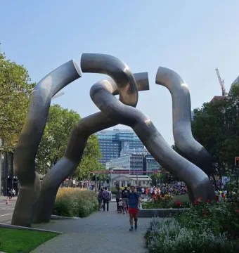 Intertwined shiny metal tube sculpture in a Berlin plaza with people and buildings nearby.