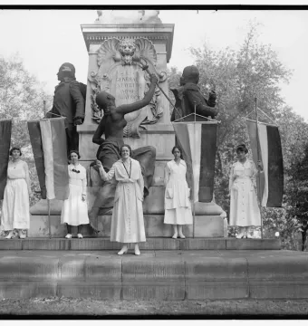 Women standing on the base of the Marquis de Lafayette Statue with suffrage flags in front of bronze figures and busts.