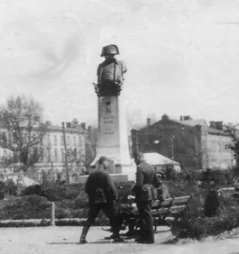 Monument to Napoleon Bonaparte on a tall plinth in a park, with people sitting and walking nearby.